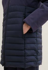 Granatowa kurtka puchowa z poziomym pikowaniem, bocznymi kieszeniami na zamek i gładką górną warstwą. Charakteryzuje się luźnym fasonem i trwałą konstrukcją.