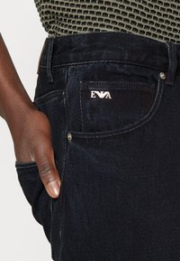Mörka denimjeans med en silverfärgad logotyp på framlappen, med synlig textur och sömnadsdetaljer. Hand placerad i fickan.
