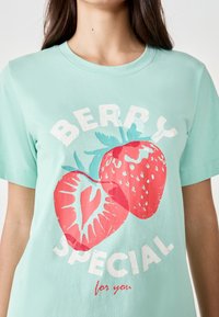 T-shirt verde menta con una grafica di due fragole rosse e testo bianco che recita "BERRY SPECIAL per te." Materiale in cotone morbido.