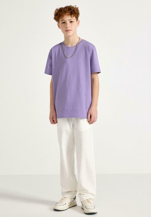 Giovane dai capelli rossi con indosso una maglietta lavanda, catena d'argento, pantaloni bianchi a gamba larga e sneakers color crema, in piedi su uno sfondo uniforme.