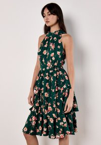 Robe sans manches à motif floral avec un fond vert, un col haut et des couches froncées. Fabriquée en tissu léger.