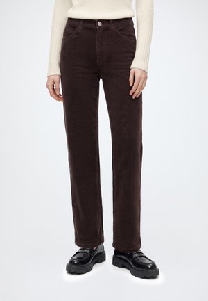 Vrouw draagt bruine corduroy broek met rechte pijpen, crème geribd langarmshirt en zwarte chunky loafers tegen een effen achtergrond.