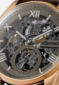 Montre squelette avec un boîtier en bronze, des engrenages noirs complexes, des accents dorés et des chiffres romains ; présente un cadran argenté texturé et un mouvement visible.