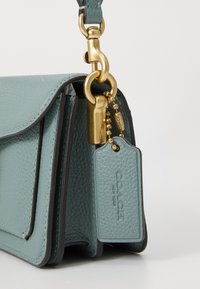 Teal leren handtas met een gestructureerd oppervlak, gouden hardware en een bagagelabel met de merknaam. Compacte, rechthoekige vorm.
