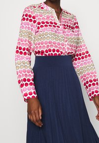 Blus med lång ärm i en rosa tyg med stora flerfärgade cirklar, framknappar och klassisk krage, i kombination med en marinblå plisserad kjol.