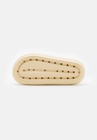 Krämfärgad gummiskosula med ett texturerat zigzagmönster, cirkulära hål för ventilation och släta kanter. Har storleksmarkeringar.