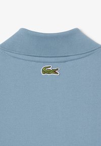 Jasnoniebieska koszulka polo z teksturowanej tkaniny i płaskim kołnierzykiem. Na plecach znajduje się wyhaftowane zielone logo krokodyla.