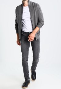 Man som bär en grå zip-up jacka över en vit t-shirt, mörkgrå skinny jeans och svarta skor som går mot en vit bakgrund.