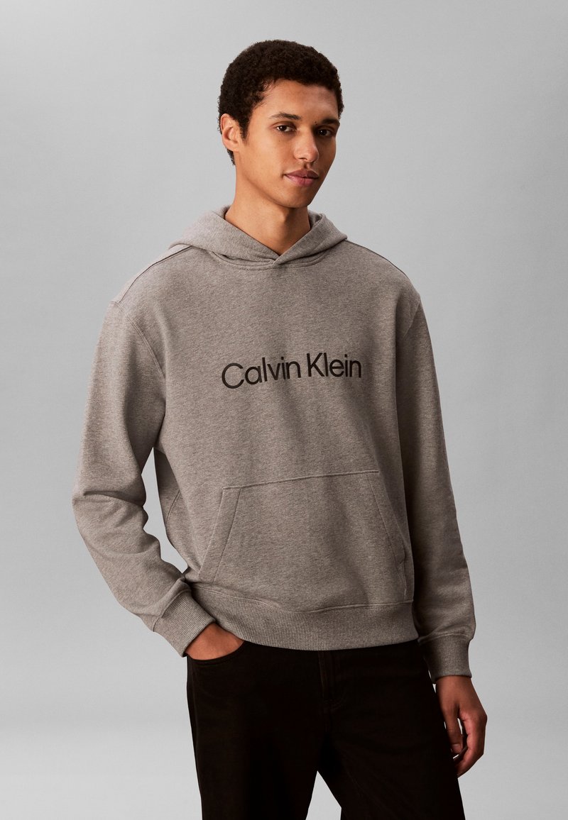 Ung mann iført en grå Calvin Klein hettegenser med frontlomme og svarte bukser, står mot en ensfarget grå bakgrunn.