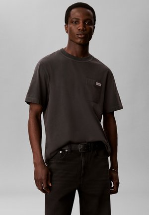 Giovane uomo che indossa una t-shirt grigio scuro a maniche corte con tasca frontale e jeans neri, in piedi davanti a uno sfondo grigio chiaro uniforme.