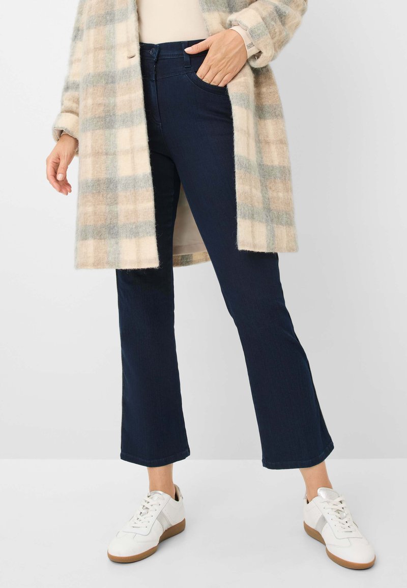 Jeans évasés bleu marine, associés à un manteau à carreaux beige et blanc et des baskets blanches avec des semelles brunes. Texture denim lisse.