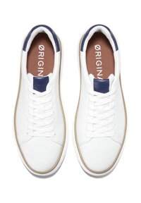 Pár bílých nízkých tenisek s námořnicky modrými patkami a stélkami označenými "ORIGINS", bílými tkaničkami a béžovým lemem kolem podrážky.