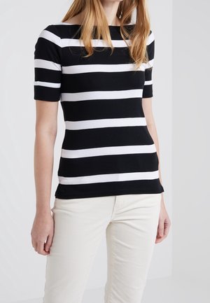 Femme portant un haut à manches courtes rayé horizontalement noir et blanc et un pantalon de couleur claire, debout devant un fond clair uni.