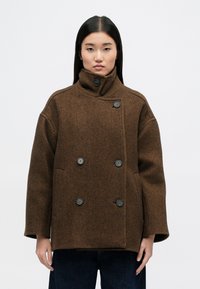 Καφέ διπλόχρωμο παλτό με ψηλό γιακά, φτιαγμένο από ύφασμα με σχέδιο ψαροκόκαλο. Διαθέτει μεγάλους σκούρους κουμπιά και πλαϊνές τσέπες.