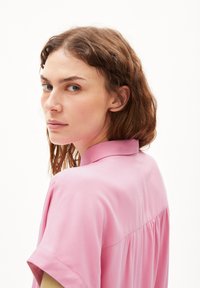 Pink shirt with a classic collar and short sleeves. Features a smooth texture and gathered detail at the back. Model looks over shoulder.
