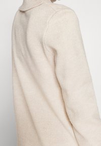 Maglione lavorato a maglia beige con colletto alto, caratterizzato da una texture morbida e una vestibilità rilassata. I dettagli delle cuciture in vista arricchiscono il design.