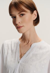 Žena s kratkom smešnom kosom koja nosi bijelu vezenku i zlatni privjesak na ogrlici, ispred jednolične pozadine.