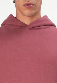 Bordo spalvos džemperis su minkšta tekstūra, su subtiliu prekės ženklo logotipu įspaustu priekyje. Jame yra standartinė kengūros tipo kišenė ir suveržimo kapučinas.