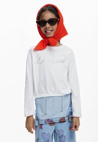Weißes, verkürztes Sweatshirt mit Strassschriftzug, kombiniert mit Jeansrömern mit Herzmuster, accessorized mit einem leuchtend orangefarbenen Schal und Sonnenbrille.
