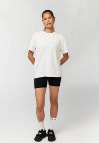 Frau steht mit den Händen auf dem Rücken, trägt ein weißes T-Shirt, schwarze Shorts, weiße Socken und schwarze Turnschuhe vor einem schlichten Hintergrund.