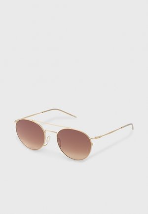 Lunettes de soleil fines à monture métallique dorée, avec verres ronds marron dégradés et plaquettes de nez réglables sur fond blanc.