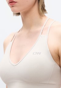 Νεαρή γυναίκα που φοράει μπεζ αθλητικό σουτιέν με διπλά λουριά και μικρό λογότυπο ICANIWILL στο στήθος.