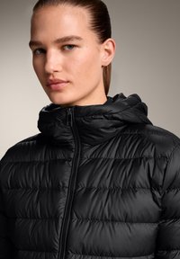Črna puhovka s kapuco, quiltana, z zadrgo na sprednjem delu in horizontalnim šivom. Gladka, sijoča tekstura z minimalističnim dizajnom.