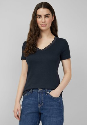 Jonge vrouw met lang golvend haar, gekleed in een marineblauw V-hals shirt met korte mouwen en kantafwerking, en een blauwe spijkerbroek, met haar hand in de zak.