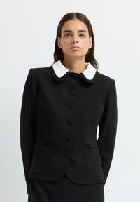 Ung kvinna som bär en svart knappjacka med en vit rundad krage, stående mot en enfärgad ljus bakgrund.