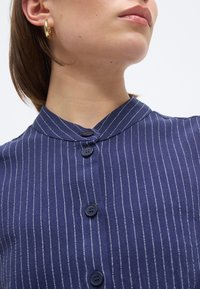 Γυναίκα που φοράει μπλε σακάκι με κάθετες λευκές ρίγες και χρυσό σκουλαρίκι κρίκο, με το κεφάλι ελαφρώς γυρισμένο προς τα πλάγια.