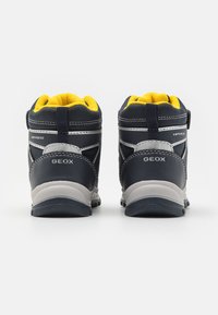 Botas impermeables para niños en color marino y gris, con acabado texturizado, cuello acolchado amarillo y detalles reflectantes. Marca Geox visible.