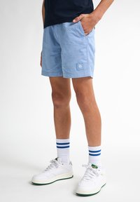 Ljust blå shorts gjorda av mjukt tyg med sidofickor och en fyrkantig logotyp. I kombination med vita sneakers och randiga sportstrumpor.