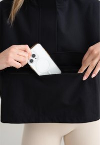 Crni skraćeni dukserica s patentnim zatvaračem i džepom. Ruka stavlja bijeli telefon s kružnim magnetom u džep. Glatka tekstura tkanine.