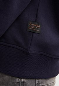 Sudadera azul marino con dobladillo y puños acanalados; incluye una etiqueta negra con "Petrol Ind" y otro texto; material de mezcla de algodón.