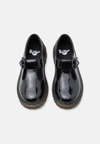 Chaussures Mary Jane Dr. Martens en cuir verni noir avec brides en T, boucles en forme de cœur, couture jaune sur les semelles et design ajouré à l'avant.