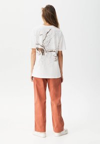 Camiseta blanca de algodón con un patrón abstracto marrón en la parte posterior, combinada con pantalones sueltos de color óxido y zapatillas blancas.