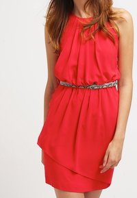 Femme portant une robe rouge sans manches avec des détails plissés et une ceinture à motif serpent, se tenant devant un fond blanc uni.