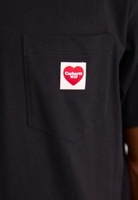 Black short-sleeve T-shirt with a chest pocket; features a square label with red heart and "Carhartt WIP" text in white. Classic cotton fabric.