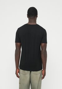 Muž stojaci chrbtom ku kamere, oblečený v jednoduchej čiernej tričku s krátkym rukávom a svetlých opotrebovaných džínsach na bielom pozadí.