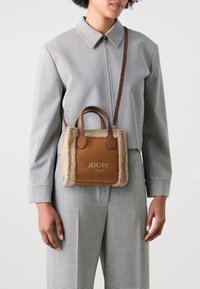 Donna che indossa un completo a scacchi bianco e nero, mentre porta una borsa a tracolla in shearling marrone con il logo "JOOP! JEANS".