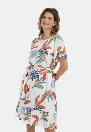 Donna sorridente che indossa un vestito bianco al ginocchio con stampe floreali tropicali colorate, maniche arricciate e cintura annodata in vita.
