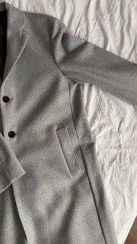 Svetlo sivi volneni plašč z oblikovalskim krojem, z zarezanim ovratnikom, dvema gumboma in enim stranskim žepom. Gladek tekstura s poliranim videzom.