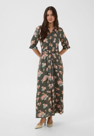 Une femme porte une robe midi vert foncé à imprimé floral, nouée à la taille et dotée de manches trois-quarts, avec des chaussures à talons beiges.