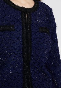 Námořnický modrý texturovaný kardigan se vzorem ve tvaru zig-zag, s černým lemem a dvěma náprsními kapsami. Materiál má jemný lesk.