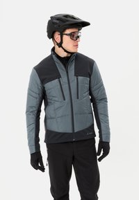 Giacca isolante grigia e nera, con tasche con zip, colletto alto e tessuto flessibile. Indossata con un casco nero e guanti.
