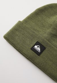 Groene gebreide beanie van zachte, gestructureerde stof. Voorzien van een zwart vierkant label met een wit logo aan de voorkant.