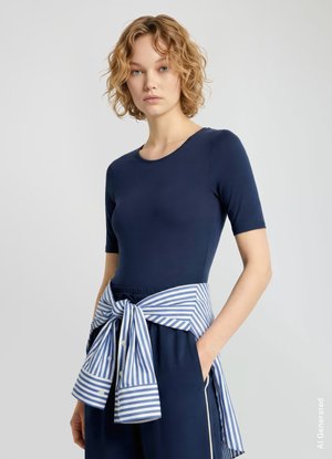 Donna con capelli corti e ricci che indossa una maglia aderente a maniche corte blu navy e pantaloni blu navy con una camicia a righe legata intorno alla vita.
