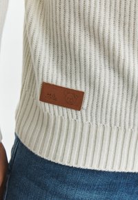 Pulover tricotat cu nervuri crem, cu o etichetă din piele maro ce prezintă un design cu urs, purtat peste blugi albaștri. Textura pare moale și confortabilă.