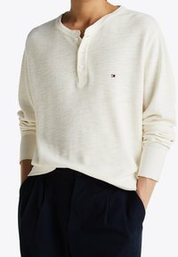 White waffle-knit long sleeve henley shirt with three buttons, featuring a small logo in blue, red, and white on the chest.
