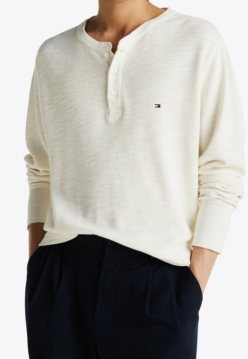 White waffle-knit long sleeve henley shirt with three buttons, featuring a small logo in blue, red, and white on the chest.